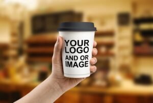 Hand holding a white paper cup with a black lid featuring placeholder text “YOUR LOGO AND OR IMAGE” in a blurred café setting.